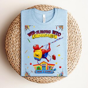 Pu&ograve; includere: Maglietta azzurra con grafica di Spider-Man che si lancia verso una scuola. Il testo recita "Web-Slinging Into Kindergarten" e "Here I Come!". La maglietta &egrave; presentata su una superficie rotonda e intrecciata.