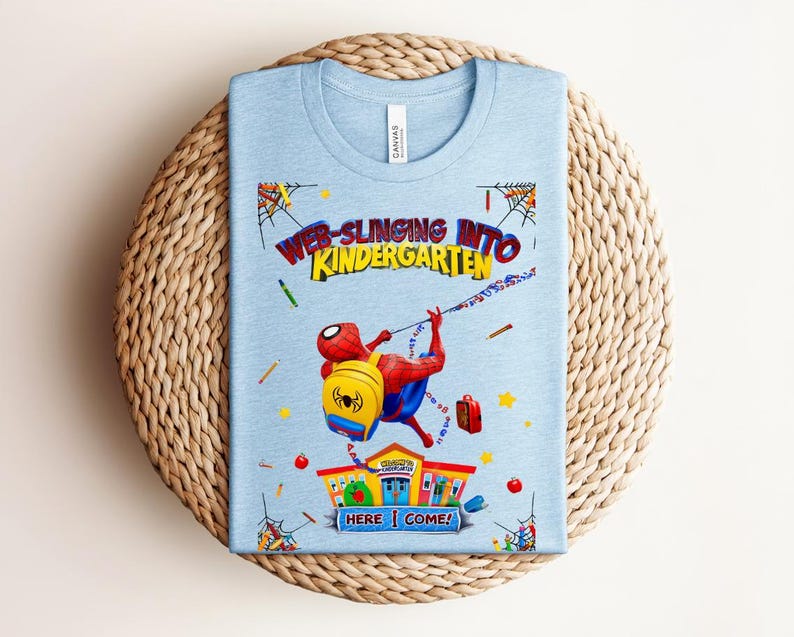 Pu&ograve; includere: Maglietta azzurra con grafica di Spider-Man che si lancia verso una scuola. Il testo recita "Web-Slinging Into Kindergarten" e "Here I Come!". La maglietta &egrave; presentata su una superficie rotonda e intrecciata.