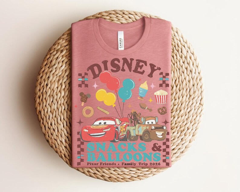 May include: A dusty rose t-shirt featuring a graphic design with the word "DISNEY" in brown, along with colorful balloons, cartoon cars, and food icons. The text "SNACKS & BALLOONS" is also displayed. The shirt is on a woven basket.