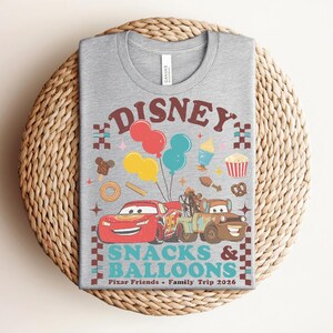 May include: A gray t-shirt featuring a colorful graphic design with the text "Disney Snacks & Balloons". The design includes cartoon cars, balloons, and food icons. The shirt is a crew neck and is made of a soft material.