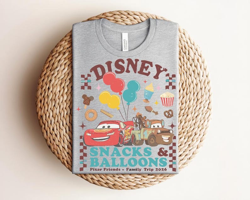 May include: A gray t-shirt featuring a colorful graphic design with the text "Disney Snacks & Balloons". The design includes cartoon cars, balloons, and food icons. The shirt is a crew neck and is made of a soft material.