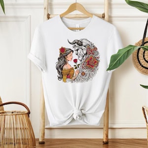 May include: White t-shirt featuring a colorful illustration of a woman and a beast, with a red rose. The design includes floral and ornamental details. The woman is wearing a yellow dress and the beast has horns.
