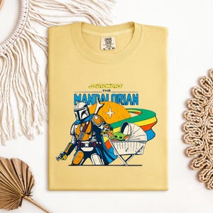 May include: A folded, light yellow t-shirt featuring a graphic from "The Mandalorian." The design includes the Mandalorian, Grogu in a floating bassinet, and the text "Star Wars The Mandalorian" in blue and orange. The shirt has a Comfort Colors tag.