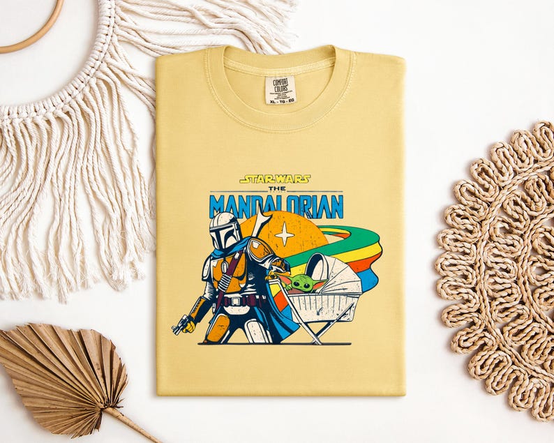 May include: A folded, light yellow t-shirt featuring a graphic from "The Mandalorian." The design includes the Mandalorian, Grogu in a floating bassinet, and the text "Star Wars The Mandalorian" in blue and orange. The shirt has a Comfort Colors tag.