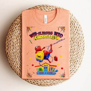 Pu&ograve; includere: T-shirt color pesca con una grafica di Spider-Man che si lancia verso la scuola materna. Il design include il testo "Web-Slinging Into Kindergarten" e "Here I Come!" con illustrazioni a tema scolastico.