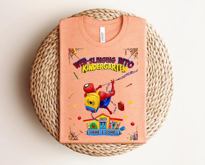 Pu&ograve; includere: T-shirt color pesca con una grafica di Spider-Man che si lancia verso la scuola materna. Il design include il testo "Web-Slinging Into Kindergarten" e "Here I Come!" con illustrazioni a tema scolastico.