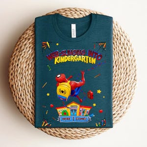 Pu&ograve; includere: T-shirt verde acqua con Spider-Man e la scritta "WEB-SLINGING INTO KINDERGARTEN". Il disegno include una scuola, uno zaino e delle stelle. La maglietta &egrave; su una superficie circolare intrecciata.