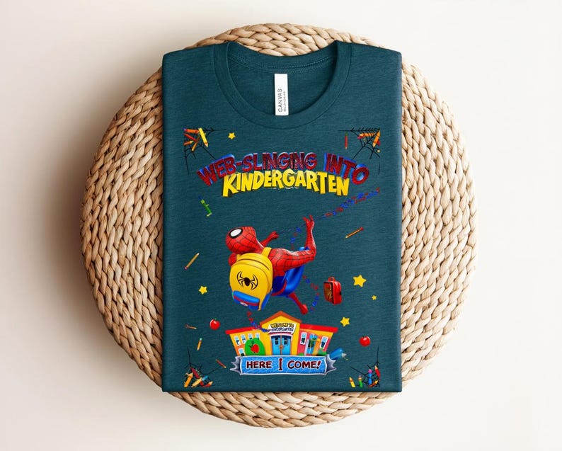 Pu&ograve; includere: T-shirt verde acqua con Spider-Man e la scritta "WEB-SLINGING INTO KINDERGARTEN". Il disegno include una scuola, uno zaino e delle stelle. La maglietta &egrave; su una superficie circolare intrecciata.