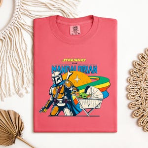 May include: A coral-colored t-shirt featuring a graphic from Star Wars: The Mandalorian. The design includes the Mandalorian, a baby Yoda character in a bassinet, and the show's logo. The shirt is a crew neck style.