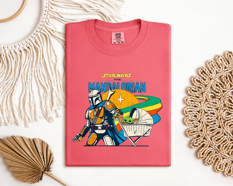 May include: A coral-colored t-shirt featuring a graphic from Star Wars: The Mandalorian. The design includes the Mandalorian, a baby Yoda character in a bassinet, and the show's logo. The shirt is a crew neck style.
