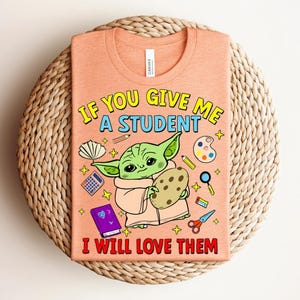 May include: Peach-coloured t-shirt with a Grogu-like cartoon character holding a cookie. The shirt displays the text "IF YOU GIVE ME A STUDENT I WILL LOVE THEM" in yellow and red. School-related illustrations surround the character.