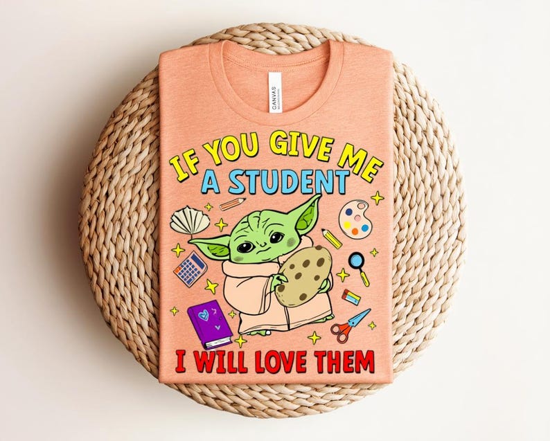 May include: Peach-coloured t-shirt with a Grogu-like cartoon character holding a cookie. The shirt displays the text "IF YOU GIVE ME A STUDENT I WILL LOVE THEM" in yellow and red. School-related illustrations surround the character.