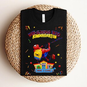 Pu&ograve; includere: T-shirt nera con una grafica colorata di Spider-Man che si lancia verso una scuola. Il testo recita "Web-Slinging Into Kindergarten" con "Here I Come!" sotto. Il design include stelle, matite e mele.