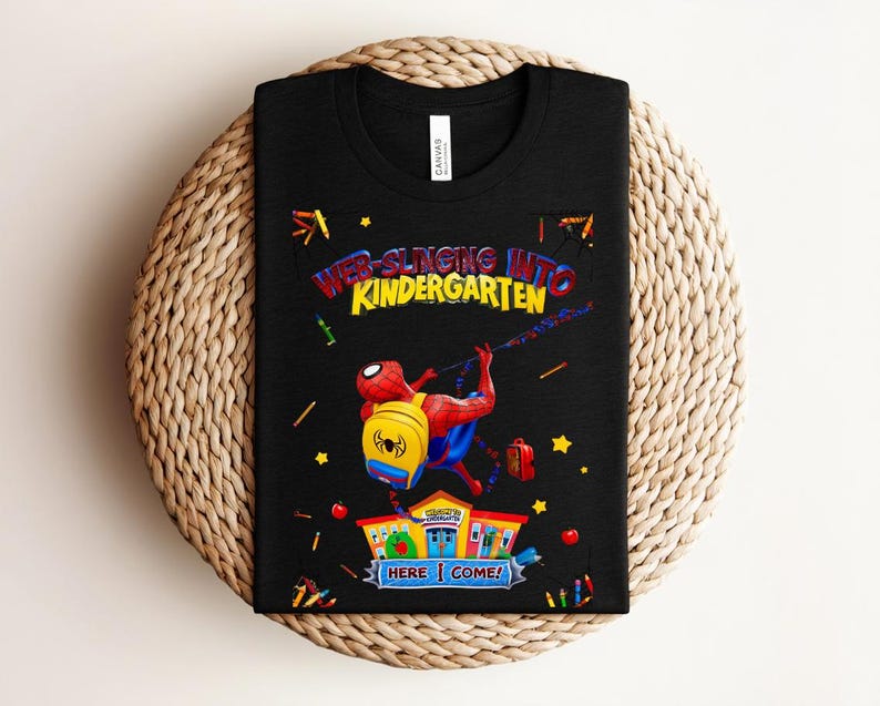 Pu&ograve; includere: T-shirt nera con una grafica colorata di Spider-Man che si lancia verso una scuola. Il testo recita "Web-Slinging Into Kindergarten" con "Here I Come!" sotto. Il design include stelle, matite e mele.