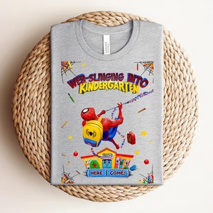 Pu&ograve; includere: Una maglietta grigia con una grafica colorata di Spider-Man che si lancia verso una scuola. Il testo recita "Web-Slinging Into Kindergarten" con l'aggiunta di "Here I Come!" Il design include uno zaino, materiale scolastico e una scuola.