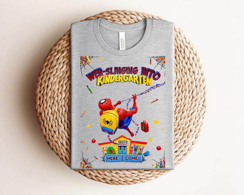 Pu&ograve; includere: Una maglietta grigia con una grafica colorata di Spider-Man che si lancia verso una scuola. Il testo recita "Web-Slinging Into Kindergarten" con l'aggiunta di "Here I Come!" Il design include uno zaino, materiale scolastico e una scuola.