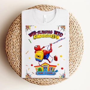 Pu&ograve; includere: T-shirt bianca con una grafica colorata di Spider-Man che si lancia verso la scuola materna. Il design include il testo "WEB-SLINGING INTO KINDERGARTEN" e "HERE I COME!" con illustrazioni a tema scolastico.