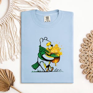 May include: Light blue t-shirt featuring a cartoon bear wearing a green scarf and holding a pot of yellow flowers. The bear is sitting on green grass. The shirt has a Comfort Colors tag.
