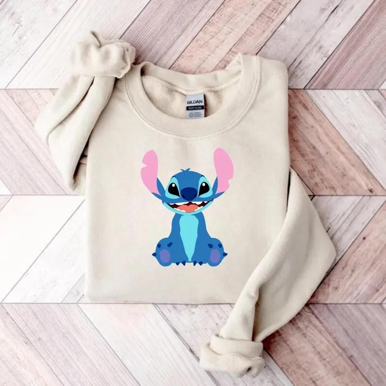May include: A beige crewneck sweatshirt featuring a cartoon image of Stitch from Lilo & Stitch. The character is depicted in shades of blue with pink ears and a smiling expression. The sweatshirt is folded on a wooden surface.