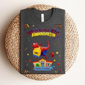 Pu&ograve; includere: T-shirt grigio scuro con una grafica colorata di Spider-Man e la scritta "WEB-SLINGING INTO KINDERGARTEN". Il design include un edificio scolastico, stelle e materiale scolastico. Un design divertente per bambini.
