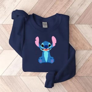 May include: Navy blue sweatshirt featuring a cartoon image of Stitch from Lilo & Stitch. The graphic shows Stitch in shades of blue with pink ears and a wide smile. The sweatshirt is made of a soft material.