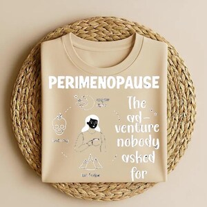 Op de afbeelding: Een beige t-shirt met het woord "PERIMENOPAUSE" in het wit. Het shirt heeft een afbeelding met een schedel, maan, vrouw en bergen, en de tekst "The adventure nobody asked for."