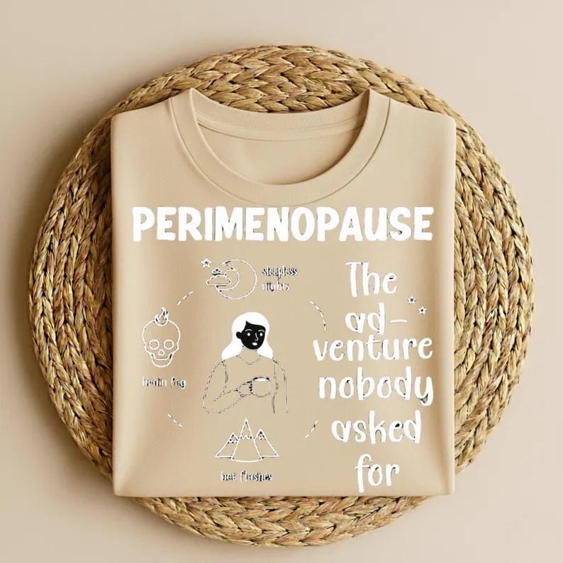 Op de afbeelding: Een beige t-shirt met het woord "PERIMENOPAUSE" in het wit. Het shirt heeft een afbeelding met een schedel, maan, vrouw en bergen, en de tekst "The adventure nobody asked for."