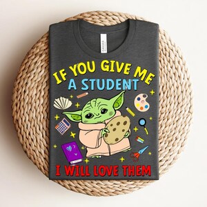 May include: Dark grey t-shirt with a cartoon character holding a cookie, surrounded by school supplies. The text reads, "IF YOU GIVE ME A STUDENT I WILL LOVE THEM" in yellow and red. The shirt is displayed on a woven surface.