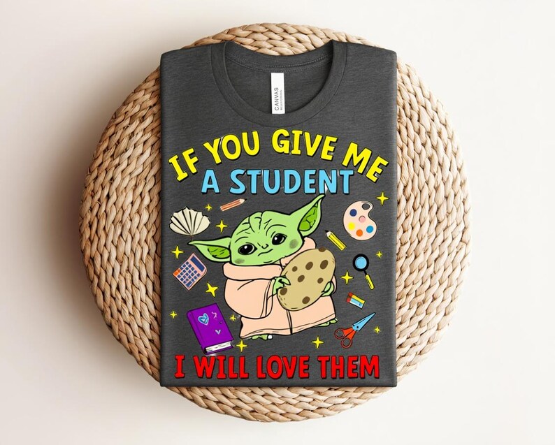 May include: Dark grey t-shirt with a cartoon character holding a cookie, surrounded by school supplies. The text reads, "IF YOU GIVE ME A STUDENT I WILL LOVE THEM" in yellow and red. The shirt is displayed on a woven surface.