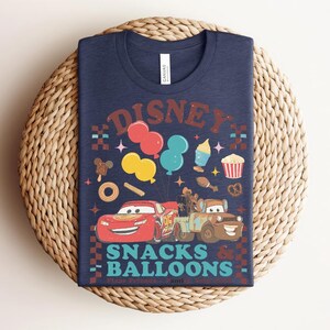 May include: Navy blue t-shirt featuring a colorful graphic with the text "Disney Snacks & Balloons." The design includes cartoon cars, balloons, and various snack illustrations. The shirt is displayed on a woven, circular surface.