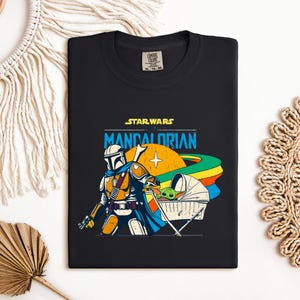 May include: Black t-shirt featuring a colorful graphic of The Mandalorian and Grogu from Star Wars. The design includes the text "STAR WARS MANDALORIAN" in yellow and blue, with a stylized illustration of the characters and a rainbow-colored planet.