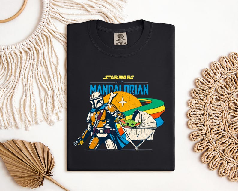 May include: Black t-shirt featuring a colorful graphic of The Mandalorian and Grogu from Star Wars. The design includes the text "STAR WARS MANDALORIAN" in yellow and blue, with a stylized illustration of the characters and a rainbow-colored planet.