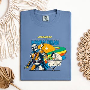 May include: A blue t-shirt featuring a graphic from "Star Wars: The Mandalorian." The design includes the Mandalorian, a baby Yoda-like character, and the text "Star Wars The Mandalorian" in yellow and blue.