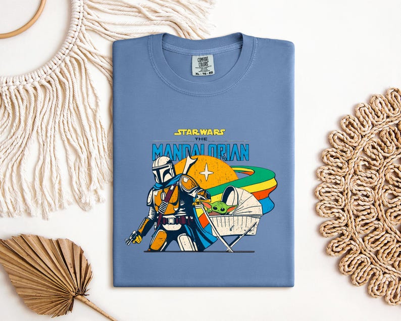 May include: A blue t-shirt featuring a graphic from "Star Wars: The Mandalorian." The design includes the Mandalorian, a baby Yoda-like character, and the text "Star Wars The Mandalorian" in yellow and blue.