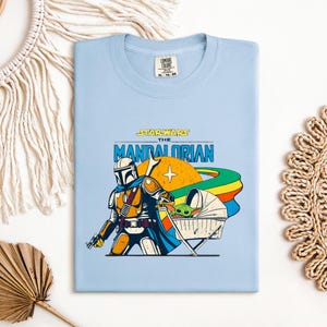 May include: Light blue t-shirt featuring a graphic of The Mandalorian from Star Wars. The design includes the Mandalorian, Grogu in a pram, and the text "STAR WARS THE MANDALORIAN". The shirt has a crew neck and a tag at the top.