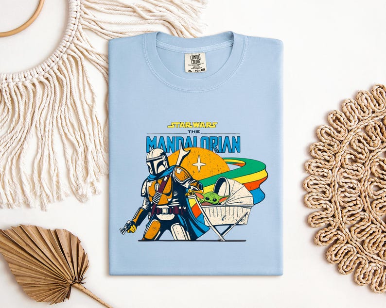 May include: Light blue t-shirt featuring a graphic of The Mandalorian from Star Wars. The design includes the Mandalorian, Grogu in a pram, and the text "STAR WARS THE MANDALORIAN". The shirt has a crew neck and a tag at the top.