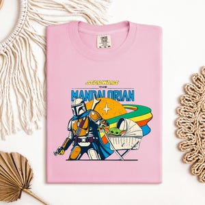 May include: A light pink t-shirt featuring a graphic of The Mandalorian and Grogu from Star Wars. The design includes the text "STAR WARS THE MANDALORIAN" in blue and yellow, with a colorful planet and Grogu in a stroller.