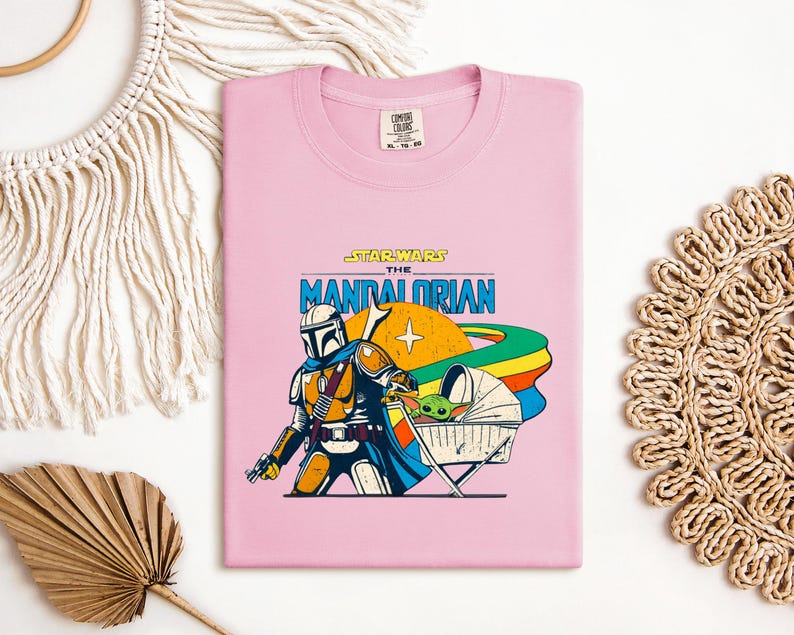 May include: A light pink t-shirt featuring a graphic of The Mandalorian and Grogu from Star Wars. The design includes the text "STAR WARS THE MANDALORIAN" in blue and yellow, with a colorful planet and Grogu in a stroller.