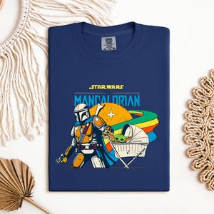 May include: Navy blue t-shirt featuring a graphic from Star Wars: The Mandalorian. The design includes the Mandalorian, Grogu in a cradle, and the text "STAR WARS THE MANDALORIAN". The artwork has orange, yellow, blue, and green accents.