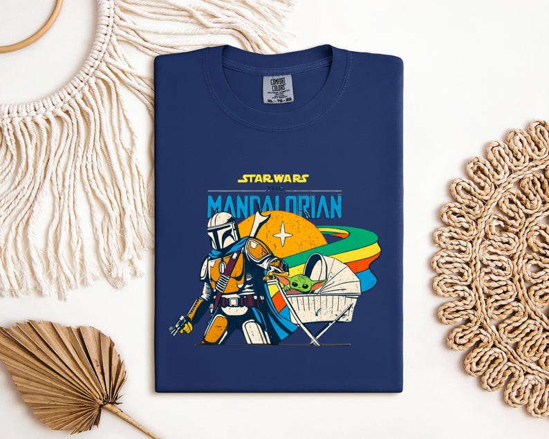 May include: Navy blue t-shirt featuring a graphic from Star Wars: The Mandalorian. The design includes the Mandalorian, Grogu in a cradle, and the text "STAR WARS THE MANDALORIAN". The artwork has orange, yellow, blue, and green accents.