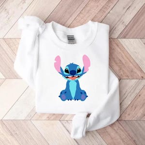 May include: White sweatshirt featuring a cartoon illustration of Stitch, a blue extraterrestrial creature with large ears and a friendly smile. The graphic is centered on the front of the long-sleeved top, which is made of a soft material.