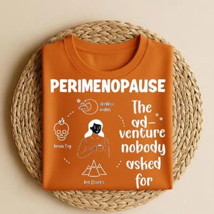 Op de afbeelding: Oranje T-shirt met het woord "PERIMENOPAUSE" in het wit. Het shirt heeft een afbeelding met illustraties van een schedel, maan, vrouw en bergen, met de tekst "brain fog", "sleepless nights" en "hot flashes". De tekst "The ad-venture nobody asked for" staat ook op het shirt.