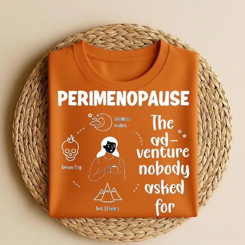 Op de afbeelding: Oranje T-shirt met het woord "PERIMENOPAUSE" in het wit. Het shirt heeft een afbeelding met illustraties van een schedel, maan, vrouw en bergen, met de tekst "brain fog", "sleepless nights" en "hot flashes". De tekst "The ad-venture nobody asked for" staat ook op het shirt.