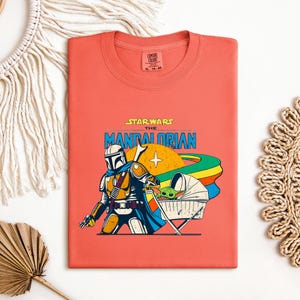 May include: Coral-colored t-shirt featuring a graphic of The Mandalorian and Grogu from Star Wars. The design includes the text "STAR WARS THE MANDALORIAN" in blue and yellow, with a retro-style illustration of the characters and a rainbow.