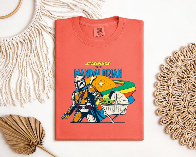 May include: Coral-colored t-shirt featuring a graphic of The Mandalorian and Grogu from Star Wars. The design includes the text "STAR WARS THE MANDALORIAN" in blue and yellow, with a retro-style illustration of the characters and a rainbow.