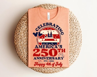 May include: Peach-coloured t-shirt with a graphic celebrating America's 250th Anniversary. The design includes a castle, stars, stripes, and the text "Happy 4th of July". The shirt is displayed on a woven, circular placemat.