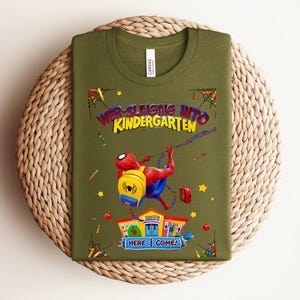 Pu&ograve; includere: T-shirt verde oliva con una grafica colorata di Spider-Man che si lancia verso una scuola. Il testo recita "Web-Slinging Into Kindergarten" con "Here I Come!" e illustrazioni scolastiche.