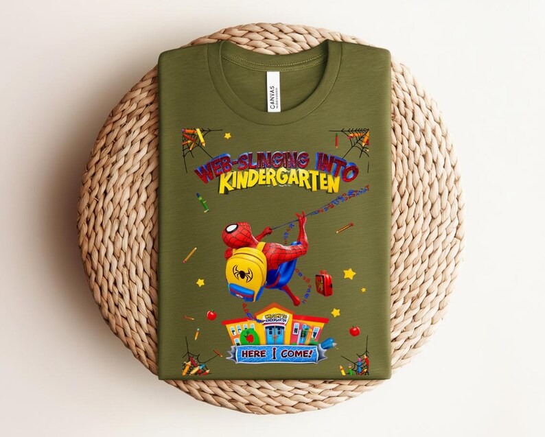 Pu&ograve; includere: T-shirt verde oliva con una grafica colorata di Spider-Man che si lancia verso una scuola. Il testo recita "Web-Slinging Into Kindergarten" con "Here I Come!" e illustrazioni scolastiche.