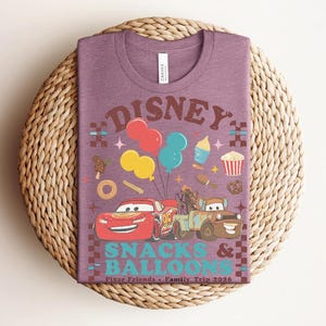 May include: A purple t-shirt featuring a graphic design with the text "Disney Snacks & Balloons". The design includes cartoon cars, balloons, and food icons. The shirt is on a woven, circular surface.
