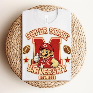May include: A white t-shirt featuring a graphic of a cartoon character wearing a red cap and varsity jacket, centered over a large red letter M. The text reads SUPER STATE UNIVERSITY EST. 1985, flanked by two brown footballs and two red stars.